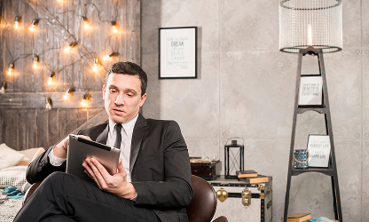 A professional man in a black suit sits in a modern, stylish lounge area while working on a digital tablet.